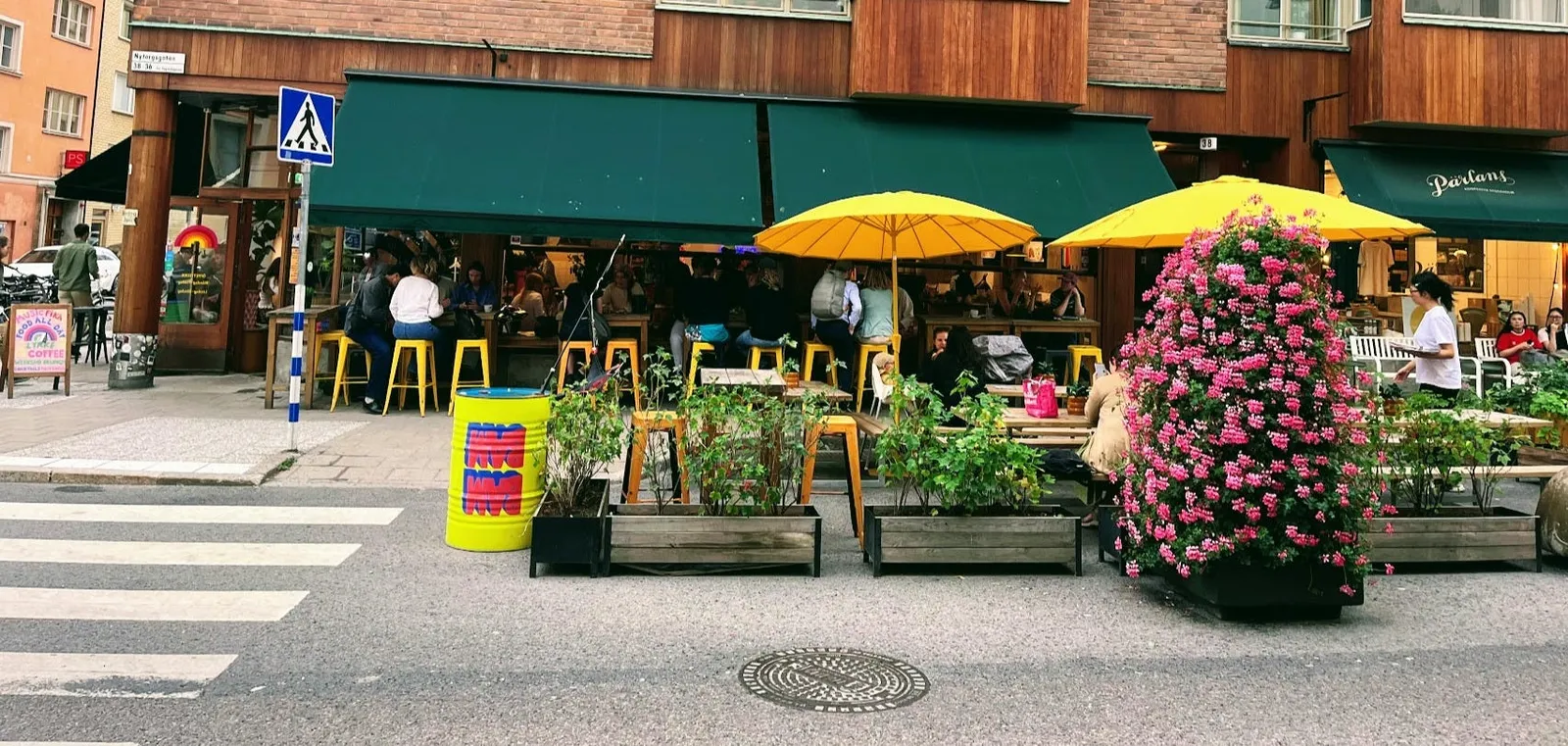 En sommargata vid Nytorget på Södermalm: fulla uteserveringar under gröna markiser och gula parasoller, med planteringslådor och ett stort torn av rosa blommor ute i körbanan.