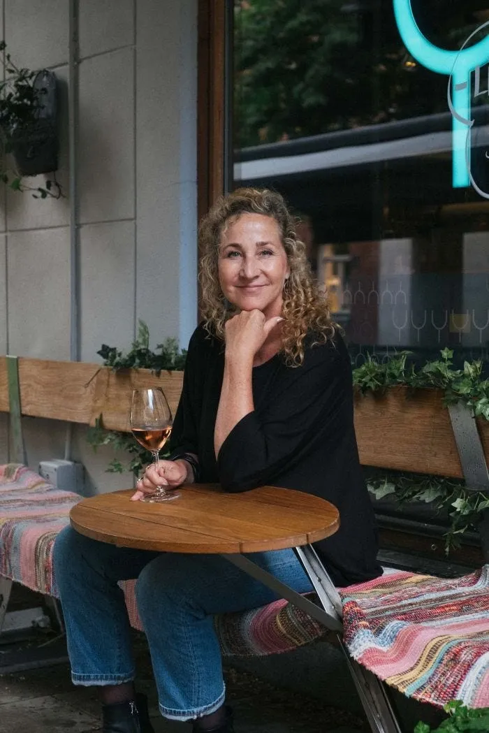 Carro Tallving, ägare till Dryck Vinbar, sitter vid ett litet runt träbord utanför vinbaren med ett glas rosé, på en bänk med färgglada trasmattor.